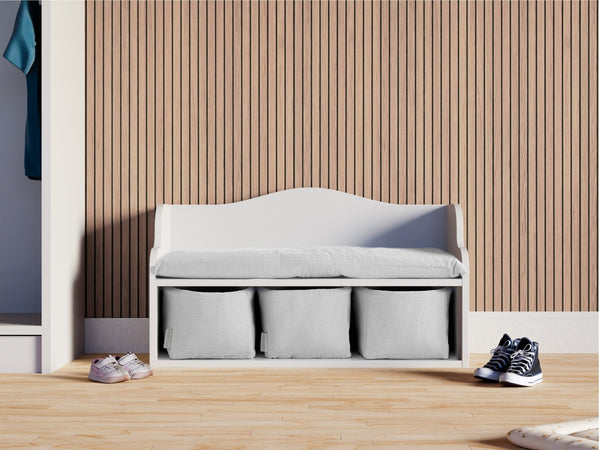 Die KraftKids Kindersitzbank weiß mit Sitzauflage steht vor einer Holzlattenwand. Drei Aufbewahrungsfächer bieten Platz für Schuhe im Kinderzimmer und machen die Bank besonders praktisch.