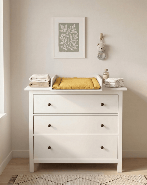 In einem minimalistischen Kinderzimmer steht eine weiße Kommode mit drei Schubladen und dem KraftKids Wickelaufsatz weiß für 108 cm breite HEMNES Kommode (Wickelfläche 50 x 70 cm), darüber eine gelbe Wickelauflage, gefaltete Handtücher und eine Flasche. Darüber hängt begrünte Kunst, durch die das Sonnenlicht fällt.