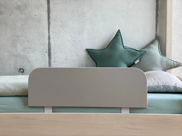 Ein minimalistisches Kinderbett mit einem grauen Kopfteil, hellem Bettzeug und zwei meergrünen Sternenkissen steht vor einer Betonwand und ist mit dem KraftKids Bettschutzgitter Rausfallschutz - grau ausgestattet, das für zusätzliche Sicherheit sorgt und leicht verstellbar ist.