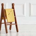 Eine kleine Holzleiter steht auf einem hellen Boden, auf dem eine KraftKids Babydecke für Babyschale Sommer aus Leinen in Gelb/Senfgelb über einer Sprosse, vor einer weißen Wand mit Zierpaneelen drapiert ist.