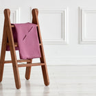 Ein hölzerner Tritthocker auf einem hellen Boden zeigt eine gefaltete KraftKids Babydecke für Babyschale Sommer Leinen magenta dunkel. Weiß getäfelte Wände bilden den Hintergrund.