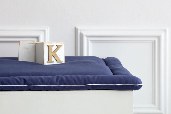 Ein Holzblock mit dem Buchstaben K ruht auf einer ordentlich gefalteten KraftKids Wickelauflage Leinen blau dunkelblau vor einer weißen Wand mit Zierleisten.