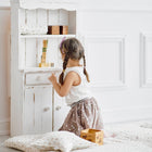 Ein kleines Kind mit geflochtenem Haar und Brille spielt mit Holzklötzen auf der Krabbeldecke Musselin Zweige braun von KraftKids, neben einem kleinen weißen Schrank. Auf dem hellen Holzboden in diesem luftigen, weißen Raum liegen Kissen und Polster.