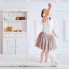 Ein kleines Kind in weißem Oberteil und gepunktetem Rock steht auf der KraftKids Krabbeldecke Musselin Zweige braun in einem hellen Raum, der sich nach oben erstreckt, mit einem kleinen weißen Regal mit Spielzeug vor einer weiß getäfelten Wand im Hintergrund.