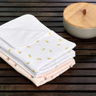 Drei fein säuberlich gefaltete Babydecken mit dezenten Blumen- und Zitronenmustern liegen auf einer dunklen Holzunterlage neben einem runden Behälter mit Holzdeckel, begleitet von der KraftKids Wickelunterlage Musselin gelbe Zitronen für den einfachen Windelwechsel unterwegs.