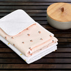 Zwei gefaltete Babydecken, eine rosa mit Blumenmuster und eine weiße mit gelbem Muster, stapeln sich auf einer dunklen Holzfläche neben der KraftKids Wickelunterlage Musselin Kirschen.