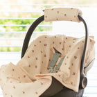 Eine KraftKids Babydecke für Babyschale Sommer Musselin Kirschen bedeckt eine leere Babyschale mit einer beigefarbenen geblümten Kopfstütze, die in einem Innenraum an einem sonnenbeschienenen Fenster mit horizontalen Jalousien angebracht ist.