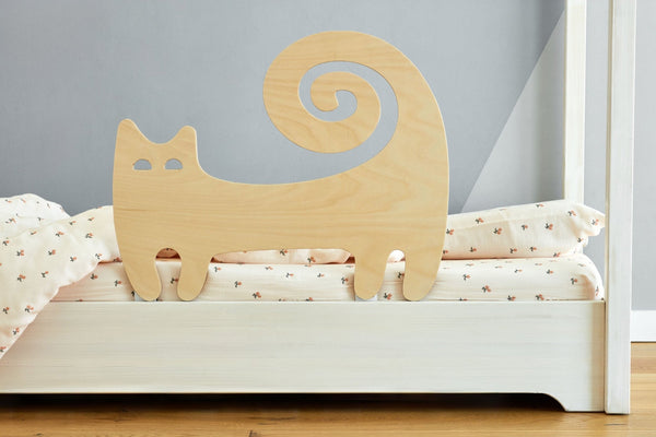 Der KraftKids Rausfallschutz aus Leimholz Katze in Form einer stilisierten Katze mit Spiralschwanz ist an einem weißen Bett mit heller Blumenbettwäsche befestigt. Eine hellgraue Wand bildet den Hintergrund.