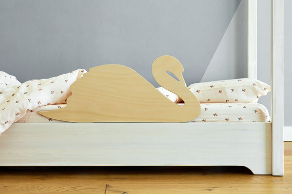 Auf einem weißen Holzbett mit gemustertem Bettzeug steht der KraftKids Rausfallschutz aus Leimholz Schwan, ein kindgerechter Bettschutz in Schwanenform aus hellem Holz, vor einer grauen Wand und einem Holzboden.