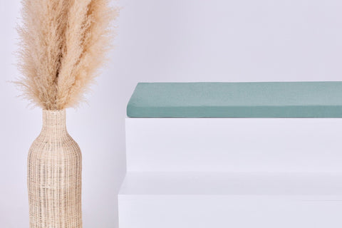 Eine hohe geflochtene Vase mit beigem Pampasgras steht neben einem weißen Stufenpodest mit einem gefalteten mintgrünen Handtuch und der KraftKids GmbH Sitzauflage für Ikea STUVA/SMÅSTAD Doppelkrepp Grün Jade vor einem schlichten hellen Hintergrund.