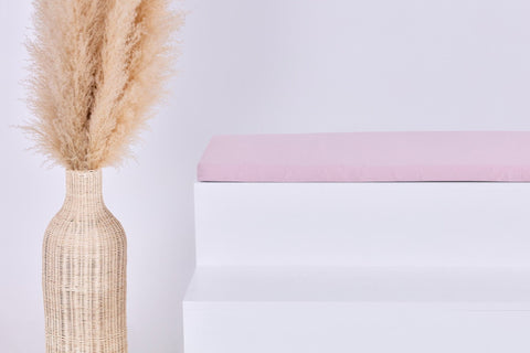 Eine hohe geflochtene Vase mit Pampasgras steht neben einem weißen Stufenpodest, auf dem die KraftKids GmbH Sitzauflage für Ikea STUVA/SMÅSTAD Doppelkrepp Rosa vor einem hellen Hintergrund steht.