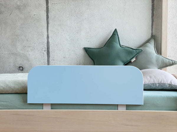 Ein modernes Kinderbett von KraftKids mit dem "Bettschutzgitter Rausfallschutz - blau hellblau", grünen sternförmigen Kissen, einem hellgrauen Kissen und Bettwäsche in sanften Pastellgrün- und Beigetönen vor einer Betonwand.