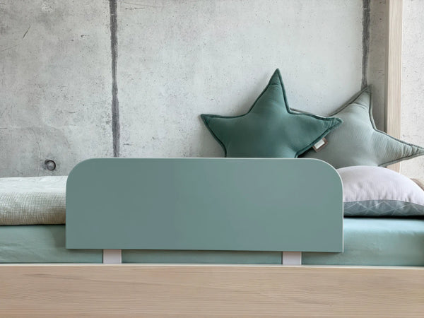 Ein minimalistisches Bett mit dem KraftKids Bettschutzgitter Rausfallschutz in blau petrolblau, gepaart mit mintgrünem Bettzeug und zwei sternförmigen Kissen vor einer Betonwand.