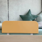 Ein minimalistisches Kinderbett mit einem hellen Holzrahmen, KraftKids Bettschutzgitter Rausfallschutz in karamellbraun, grünen sternförmigen Kissen und einem Wandhintergrund aus Beton.