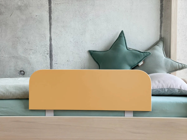 Ein minimalistisches Kinderbett mit einem hellen Holzrahmen, KraftKids Bettschutzgitter Rausfallschutz in karamellbraun, grünen sternförmigen Kissen und einem Wandhintergrund aus Beton.