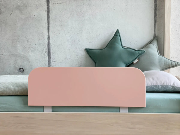 Ein minimalistisches KraftKids-Bett mit einem hellen Holzrahmen ist mit dem Bettschutzgitter Rausallschutz in Pastellrosa, grünen Sternkissen und einem weißen Kissen ausgestattet und steht vor einer kahlen Betonwand.