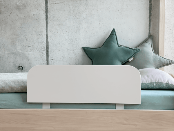 Ein minimalistisches und sicheres Kinderbett mit blassgrüner und weißer Bettwäsche, zwei großen sternförmigen Kissen und dem KraftKids Bettschutzgitter Rausfallschutz in weiß für sicheren Schlaf, vor einer hellen Betonwand.