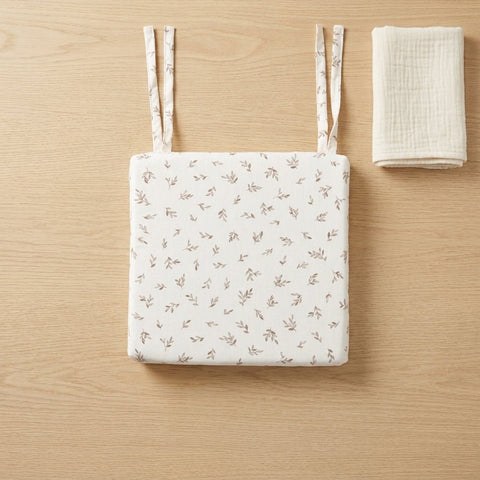 Ein KraftKids Sitzkissen für Stühle aus Musselin mit kleinen Blumendrucken und Bindebändern ruht auf hellem Holz neben einem gefalteten weißen Tuch, das einen abnehmbaren Bezug zur einfachen Reinigung aufweist.