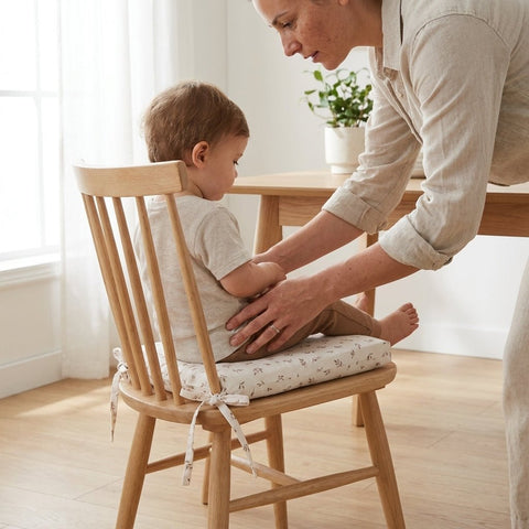 Ein Erwachsener hilft einem Kleinkind mit dem KraftKids Sitzkissen für Stühle aus Musselin in einem hellen, minimalistischen Esszimmer mit Holztisch, großem Fenster und Topfpflanze sicher auf einem gepolsterten Holzstuhl zu sitzen.