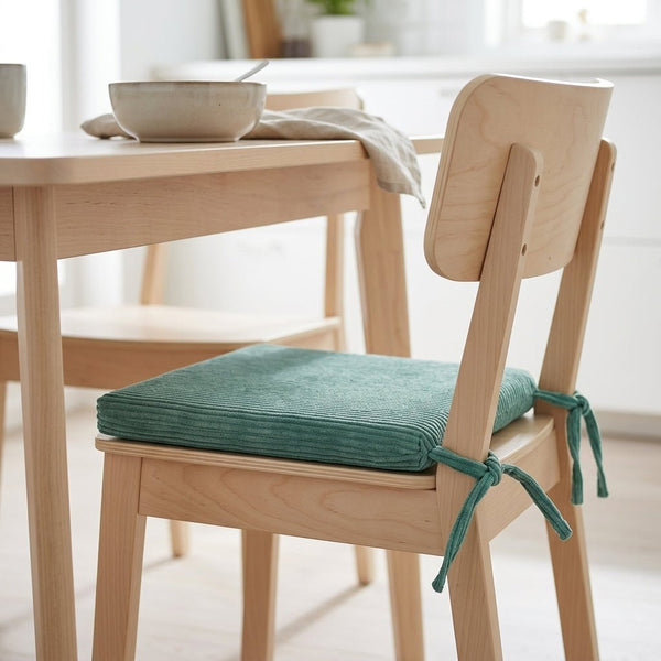 Ein heller Holzstuhl mit einem grünen KraftKids Sitzkissen für Stühle aus Breitcord ist an der Sitzfläche befestigt. Er steht neben einem passenden Tisch mit einer Schüssel und einem Geschirrtuch in einer hellen, minimalistischen Küche.