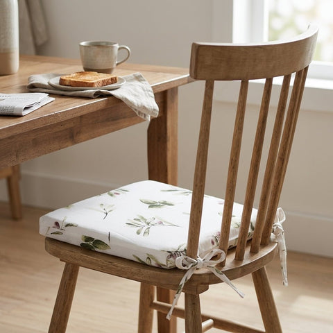 Ein Holzstuhl mit einem KraftKids Sitzkissen für Stühle aus Musselin auf der Sitzfläche sitzt neben einem Holztisch mit Tasse, Toast und Serviette in einem gemütlichen, hellen Raum. Der abnehmbare Bezug des Kissens bietet zusätzlichen Komfort und Bequemlichkeit.