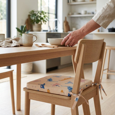 Eine Person stellt eine Schüssel auf einen hölzernen Esstisch mit Bechern, Holzutensilien und einer Serviette. Neben dem Tisch steht ein Stuhl mit dem KraftKids Sitzkissen für Stühle aus Musselin in einer hellen, modernen Küche.