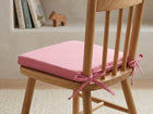 Ein Holzstuhl mit einem rosa KraftKids Sitzkissen für Stühle aus Musselin an der Rückenlehne steht auf einem hellen Teppich, während Bücher und ein kleines Holztier das Regal im Hintergrund bereichern.