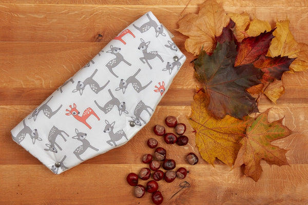 Ein KraftKids Kinderwagenmuff mit kleinen Rehkitzen in Grau und Orange auf Weiß liegt auf einer Holzoberfläche neben Herbstlaub und Kastanien.