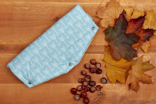 Der KraftKids Kinderwagenmuff weiße Pfeile auf Blau, ein hellblauer Fleece mit weißem Pfeilmuster und Metalldruckknöpfen, liegt auf Holz inmitten von Herbstblättern und Kastanien - ideal, um die Hände an kühlen Tagen warm zu halten.