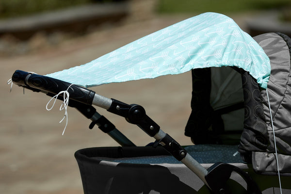 Nahaufnahme eines Kinderwagens mit dem Sonnensegel "weiße Pfeile auf Mint" von KraftKids, der leichten Schatten spendet. Der Hintergrund im Freien ist leicht verschwommen.