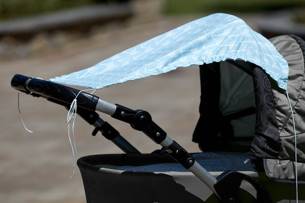 Nahaufnahme eines Kinderwagens mit dem KraftKids Sonnensegel "weiße Pfeile auf Blau" als Schattenspender drapiert. Perfekt für optimalen Sonnenschutz des Babys beim Spaziergang im Freien an einem sonnigen Tag.
