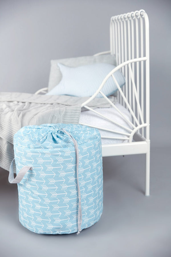 Der KraftKids Spielzeugkorb weiße Pfeile auf Blau steht auf dem Boden neben einem weißen Metallbett mit grauem Bettzeug und einem blauen Kissen und bringt Ordnung in den Raum vor grauem Hintergrund.
