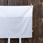 Ein Bezug für Wickeltischauflage von KraftKids mit weißen Pfeilen auf grauem Hintergrund aus 100% Baumwolle hängt vor einem rustikalen Holzzaun, an einer Leine mit Wäscheklammern befestigt.