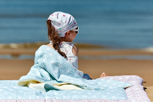 Ein junges Mädchen mit Kopftuch und Sonnenbrille sitzt auf der KraftKids Babydecke weiße Diamante auf Pastell Blau am Strand und blickt mit dem Rücken zur Kamera aufs Meer. Der Sandstrand und das ruhige blaue Wasser sind im Hintergrund zu sehen.