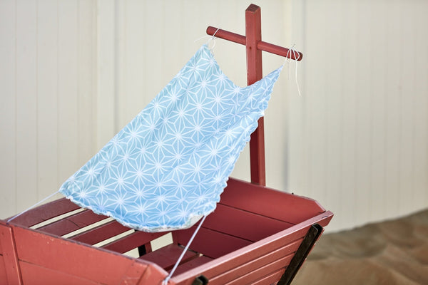 Ein KraftKids Sonnensegel "weiße Diamante auf Pastell Blau" mit weißen Rautenmustern ist über einen roten Holzwagen auf Sand drapiert, mit einem weißen Zaun im Hintergrund.