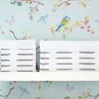 Auf einem weißen Regal steht das Körbchen graue Striche auf Weiß von KraftKids vor einer Tapete mit Vögeln, Schmetterlingen, Libellen und Blumen auf hellblauem Hintergrund.