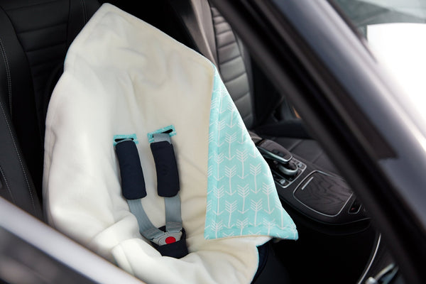Über einem leeren Autositz ist eine KraftKids "Decke für Babyschale Winter weiße Pfeile auf Blau" drapiert, mit sichtbaren schwarzen Gurtbändern und einer roten Schnalle.