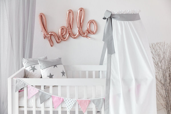 Ein weißes Babybett mit sternförmigen Kissen, einem Betthimmel Unigrau von KraftKids aus 100% Baumwolle und pastellfarbenen Wimpeln. Über der Krippe buchstabieren roségoldene Luftballons "Hallo" auf eine weiße Wand.