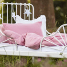 Ein weißes Metallbett mit KraftKids Bettwäscheset Musselin rosa und einem rosafarbenen Kissen steht im Freien auf einer Wiese. Am Kopfteil hängt sternförmiges Dekor inmitten von Bäumen - perfekt für erholsamen Schlaf in der Natur.