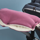 Der KraftKids Kinderwagenmuff Musselin purpur in lila und creme wird am Kinderwagengriff befestigt und bietet kuschelige Wärme für die schiebende Person. Der Hintergrund ist hellblau.