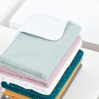 Ein ordentlich gestapeltes Set von KraftKids Musselin-Decken in Pastell- und Erdtönen ruht auf einer weißen Fläche. Obenauf liegt die "Wickelunterlage Musselin goldene Punkte auf Grün", bei der eine Ecke umgeschlagen ist und das weiße, strukturierte Futter zum Vorschein kommt.