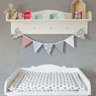 Ein KraftKids Hängeregal weiß enthält Windeln, einen Stoffhasen, eine Schnabeltasse und Dekoration über einem Wickeltisch mit Sternenmuster. Graue, blaue und rote Wimpel schmücken die graue Wand unter dem Regal.