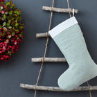 Die KraftKids Weihnachtssocke Musselin goldene Punkte auf Grün hängt auf einer rustikalen Holzleiter neben einem festlichen roten Beeren- und grünen Blätterkranz vor einem grauen Hintergrund - ideal als stilvolle Weihnachtsdeko.