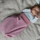 Ein Baby liegt auf einem beigen Bett, eingewickelt in eine rosa KraftKids Strampelsack Frühling Sommer Musselin rosa Wickeldecke, trägt ein weißes langärmeliges Hemd und blickt mit einem angewinkelten Arm und dem anderen am Kopf nach oben.
