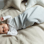 Ein Baby in weißem Outfit und KraftKids Strampelsack Frühling Sommer Musselin grau Pusteblumen liegt auf einem beigen Bett mit weichen Kissen und schaut mit erhobenem Arm auf.