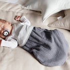 Ein Baby schläft friedlich auf einem beigen Bett, gemütlich in dem KraftKids Strampelsack Herbst Winter Musselin mit goldenen Punkten auf grau, perfekt für kühle Nächte und mit einem Schnuller in der Hand, mit sternförmigen Kissen in der Nähe.