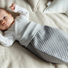 Ein Baby liegt in einem beigen Bettchen mit weißem Oberteil und grauem KraftKids Strampelsack Herbst Winter Waffel Piqué und blickt mit erhobenen Armen in die Kamera, umgeben von weichen Kissen, darunter ein sternförmiges - ideal für gemütliche Herbst- und Wintertage.