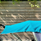 Das KraftKids Sonnensegel Musselin goldene Punkte auf Petrol beschattet einen Kinderwagen im Freien an einem Holzzaun. Sonnenlicht und Blätter werfen Schatten auf den atmungsaktiven Baumwollstoff mit goldenen Punkten.