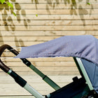 Nahaufnahme eines Kinderwagens mit einem KraftKids Sonnensegel Musselin-Verdeck in Grau mit goldenen Tupfen. Der Griff ist mit braunem Stoff umwickelt, und im Hintergrund sind die Schatten der Blätter eines Holzzauns zu sehen.