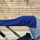 Nahaufnahme eines Kinderwagens mit dunkelblauem KraftKids Sonnensegel Musselin Verdeck, braunem Griff und hellgrauem Gestell vor Holzplanken und grünen Blättern. Das atmungsaktive Baumwollmaterial sorgt für Komfort und Schutz.